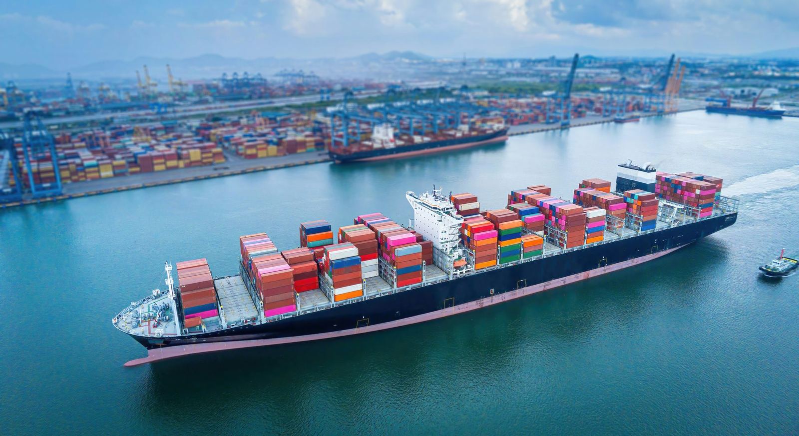 A loaded container ship moves through a harbour past a terminal stacked with containers and cranes.