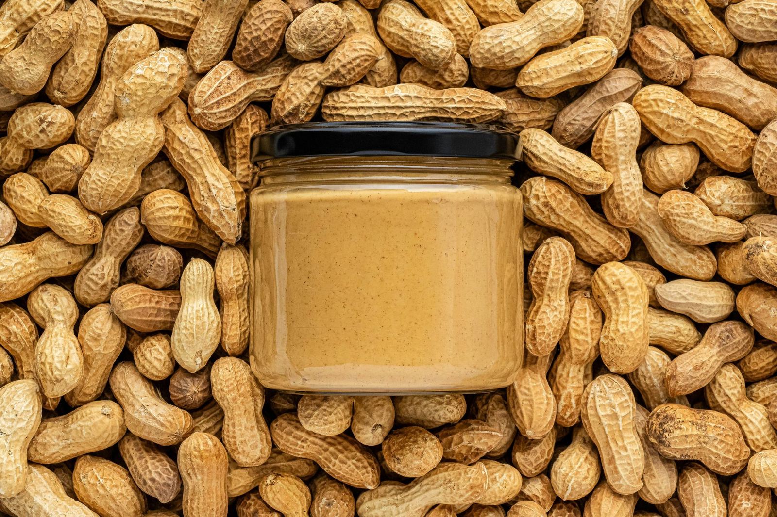 An unlabelled glass jar of peanut butter lies on a bed of peanuts in their shells.