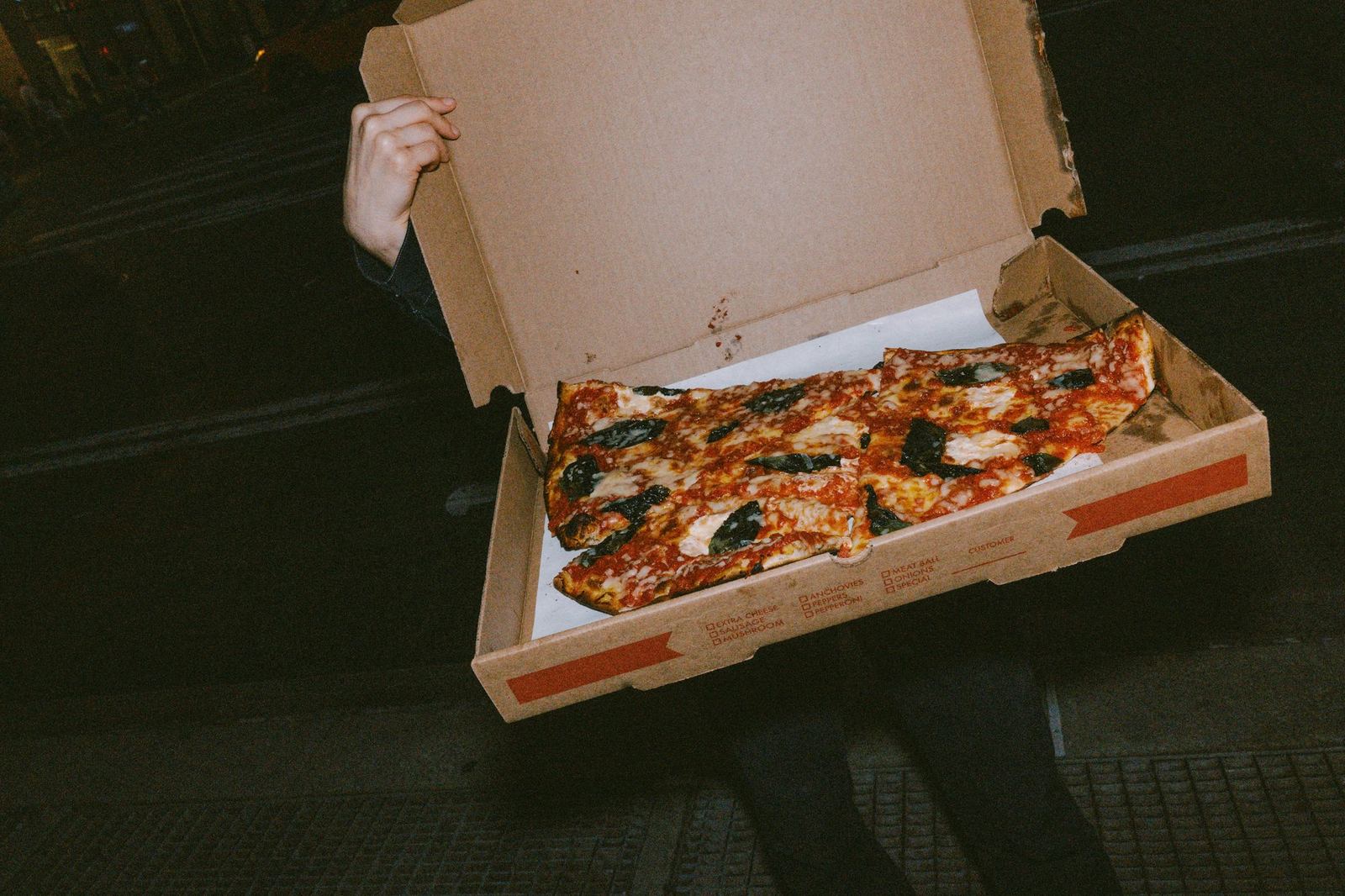 A person holds open a plain takeaway pizza box containing slices topped with tomato, cheese and basil.