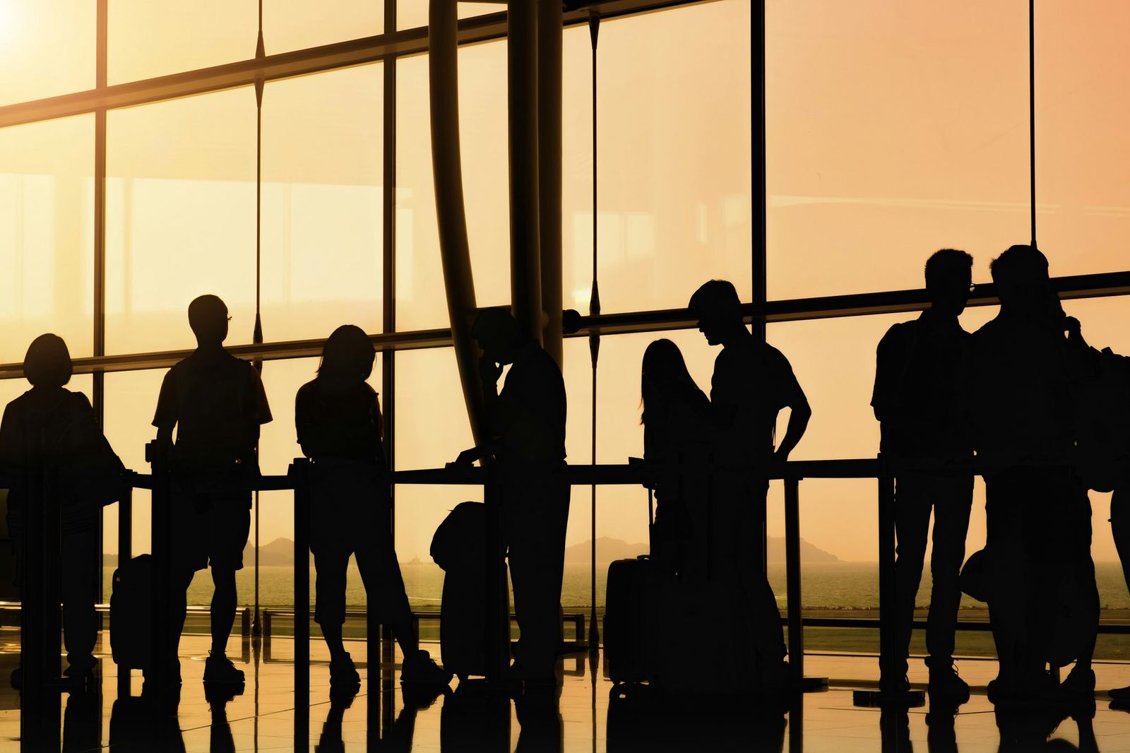 Silhouetted travellers with suitcases queue in an airport terminal in front of tall windows.