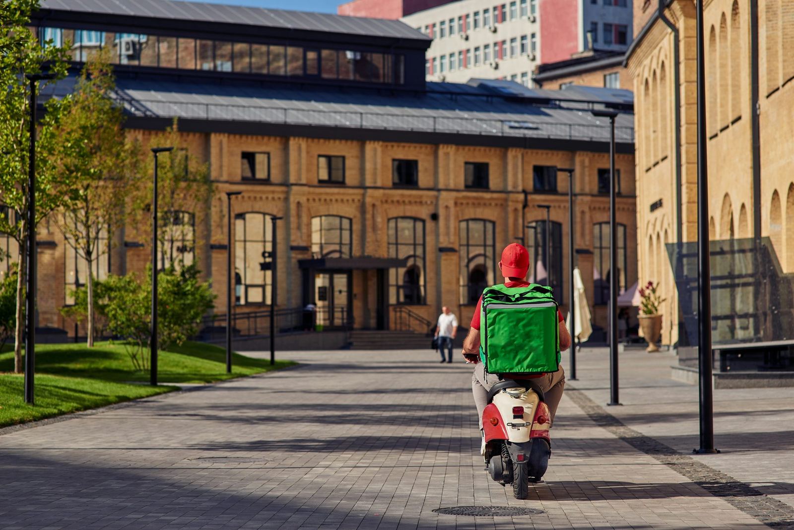 A courier with a green insulated delivery bag rides a scooter along a paved city street.